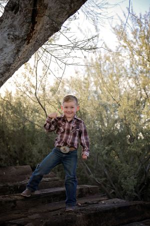 A smiling young boy in a plaid shirt, jeans and a big belt buckle poses playfully on rustic wooden steps among desert trees.