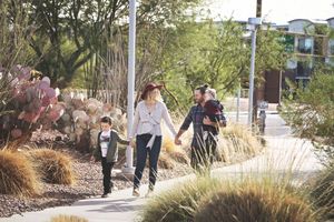 Family of four holds hands and strolls along a landscaped path with prickly pear cactus and tall grasses on a sunny day.