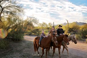 A family rides three horses along a sandy desert trail with green shrubs, mountains and a blue sky with clouds in the background.