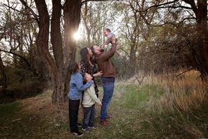 Family with two children and a lifted baby gathered beside a tree in a sunlit autumn woodland.