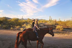 Two riders on horseback travel a dirt trail through a sunny Arizona desert landscape dotted with saguaro cacti and green shrubs.