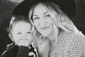 Black and white portrait of a smiling blonde woman in a wide-brim hat cuddling a toddler who chews on his fist.
