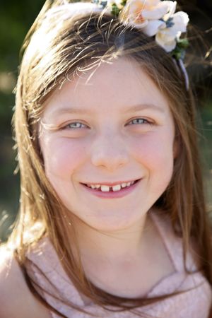 Smiling young girl with long brown hair and a white flower crown poses outdoors in soft golden sunlight.