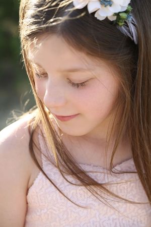 Young girl with long brown hair and a floral headband looks down with a gentle smile in warm outdoor light.