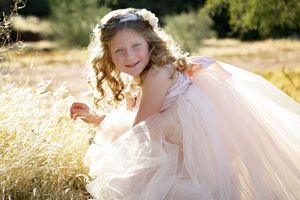Girl with curly hair and a floral headpiece wears a flowing blush tulle dress while sitting in a golden grassy field.