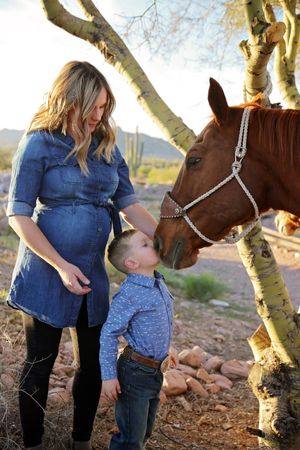 Pregnant woman in a denim dress and a young boy in a blue plaid shirt pet a chestnut horse beside a desert tree.
