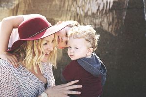 Smiling mother in a red wide-brim hat holds a curly-haired toddler while her older son leans in affectionately beside her.
