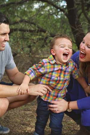 Laughing toddler in a colorful plaid shirt is held by his joyful parents while playing outdoors among trees.