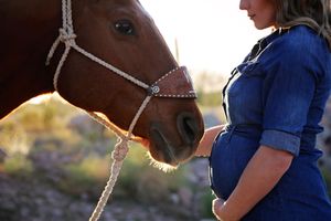 Pregnant woman in a denim dress rests her hand on her belly beside a chestnut horse in warm desert sunlight.