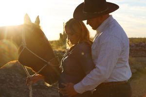 Man in a cowboy hat embraces his pregnant partner beside a chestnut horse as the golden sun sets behind them.