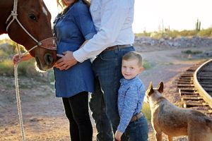 Smiling boy stands with his parents, a chestnut horse and a dog near railroad tracks in a desert at sunset.