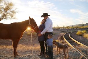 Cowboy, woman and young child stand with a brown horse and dog beside a railroad track in a sunlit Arizona desert at sunset.