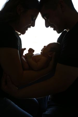 Silhouette of two parents touching foreheads while cradling their newborn baby against a bright white backlit window.