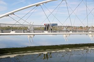 Modern white cable-stayed pedestrian bridge reflected in calm water, with two people standing on the walkway under a blue sky.