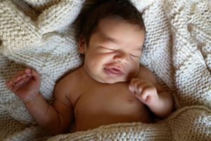 Newborn baby with dark hair sleeping peacefully and smiling slightly on a soft cream knit blanket.