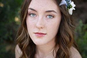 Portrait of a young woman with freckles, blue-green eyes and wavy brown hair adorned with a small floral hair accessory.