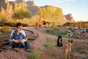 Couple in cowboy hat sitting on desert railroad tracks with two dogs and horses near rocky canyon cliffs at golden hour.