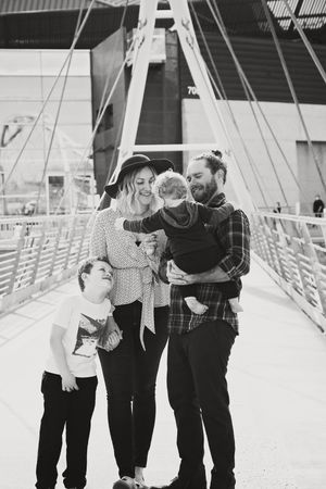 Black and white photo of a smiling family on a footbridge, with a father holding a toddler and an older boy looking up at his mom.