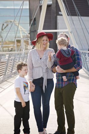 Smiling mother in a red hat holds her son's hand while the father carries a toddler on a sunlit pedestrian bridge.