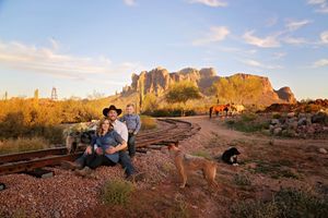 Family portrait on a desert railroad track at sunset with two dogs, horses and rugged mountain cliffs in the background.