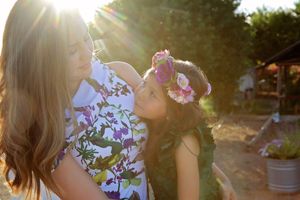 Mother and daughter embracing in warm sunlight, the girl wearing a colorful flower crown and a leafy green dress.