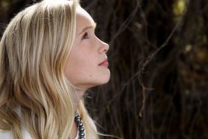 Profile portrait of a young blonde girl wearing a beaded necklace and gazing upward against a dark wooded background.