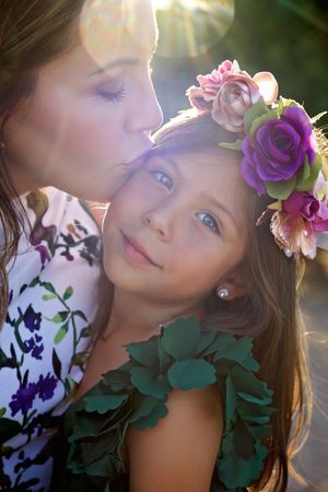 Mother kissing her smiling daughter's forehead, the girl wearing a purple and pink flower crown in soft golden light.