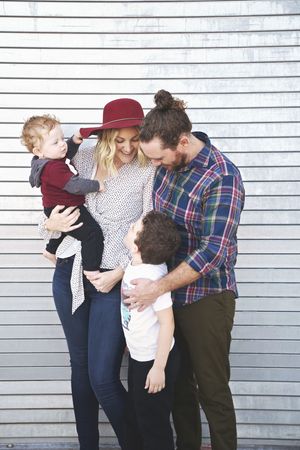 Family of four poses against a white corrugated wall, with a mother in a maroon hat holding a toddler and the father embracing an older son.