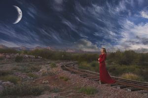 Pregnant woman in a flowing red maternity gown standing by railroad tracks under a dramatic night sky with a crescent moon.