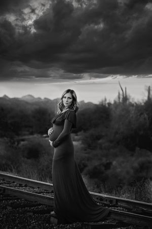 Black and white maternity portrait of a pregnant woman in a long gown standing on railroad tracks beneath a dramatic stormy desert sky.