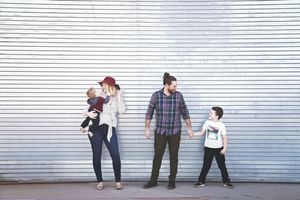 A mother holding a baby and a father holding hands with a young boy stand together in front of a white corrugated metal wall.