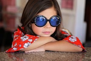 Young girl in round blue mirrored sunglasses and a red floral top rests her arms on a counter with a serious expression.