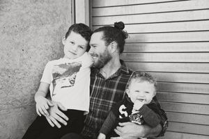 Black and white photo of a bearded father with a man bun sitting with his smiling older son and a giggling toddler on his lap.