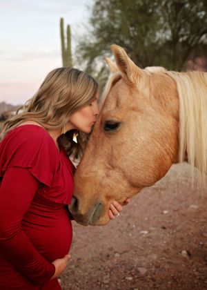 Pregnant woman in a red dress kisses the forehead of a palomino horse in a desert setting with saguaro cacti behind them.