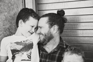 A smiling boy leans his forehead against his bearded father, who wears a man bun and plaid shirt, in a black and white family portrait.