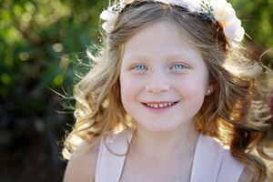 Smiling young girl with wavy blonde hair and a floral flower crown poses in warm sunlight against a leafy green background.