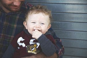 A smiling baby in a maroon shirt chews on his fist while being held by a man in a green plaid flannel shirt.