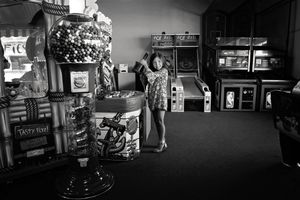 Black and white photo of a girl in a patterned dress standing beside a large gumball machine in a vintage arcade.