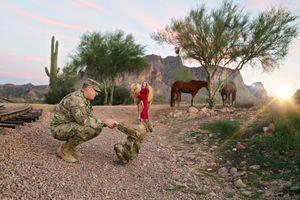 A soldier in camouflage kneels beside a child in red as horses graze in a desert landscape with saguaro cacti at sunset.