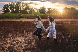 Two girls hold hands and walk across a charred field toward the glowing sunset, with trees lining the horizon.