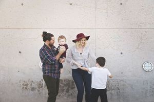 A bearded father holding a baby and a mother in a maroon hat stand against a concrete wall as a young boy walks toward them.