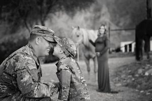 Black and white photo of a soldier father touching noses with his child in matching camouflage as a pregnant mother and horses look on.