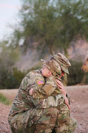 A soldier in camouflage uniform kneels and tenderly hugs a child dressed in matching military fatigues outdoors at sunset.