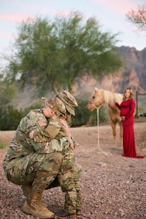 A kneeling soldier embraces a child in matching camouflage while a pregnant woman in a red gown stands with a horse behind them.