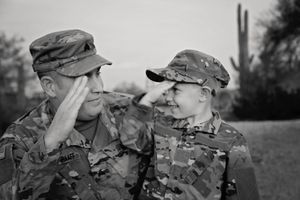 Black and white photo of a soldier and a young boy in matching camouflage uniforms saluting each other with smiles outdoors.