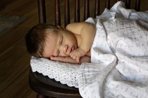 Newborn baby sleeps peacefully on a wooden chair with a lace-trimmed cushion, covered by a soft white patterned blanket.