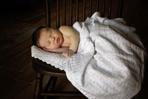 Sleeping newborn baby rests on a lace cushion atop a vintage wooden chair, draped in a white blanket against a dark background.