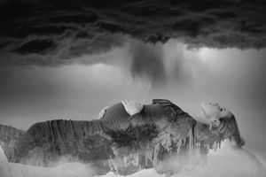 Surreal black and white maternity image of a pregnant woman lying beneath dark storm clouds in an inverted, dreamlike landscape.