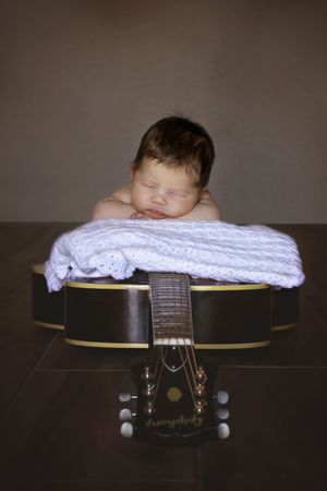 Newborn baby sleeping peacefully on a white knit blanket resting atop an acoustic guitar, with the guitar neck pointing toward the camera.
