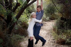 Smiling blonde girl in a white dress hugs a boy who hangs from a tree branch along a wooded dirt path outdoors.